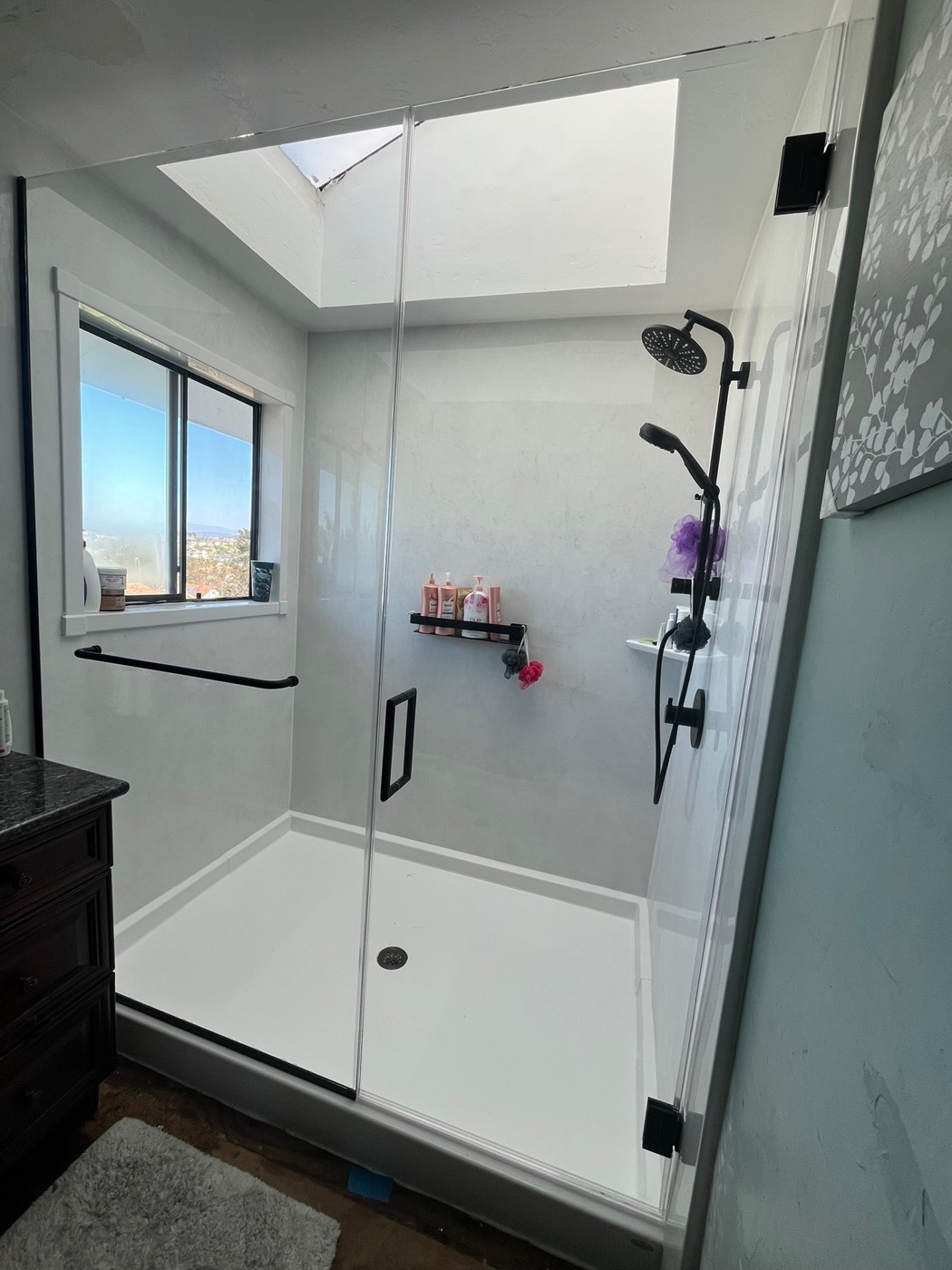 Bright walk-in marble-look stone shower with a glass door, matte-black fixtures, and a safety grab bar — a Forever Bath install in San Diego